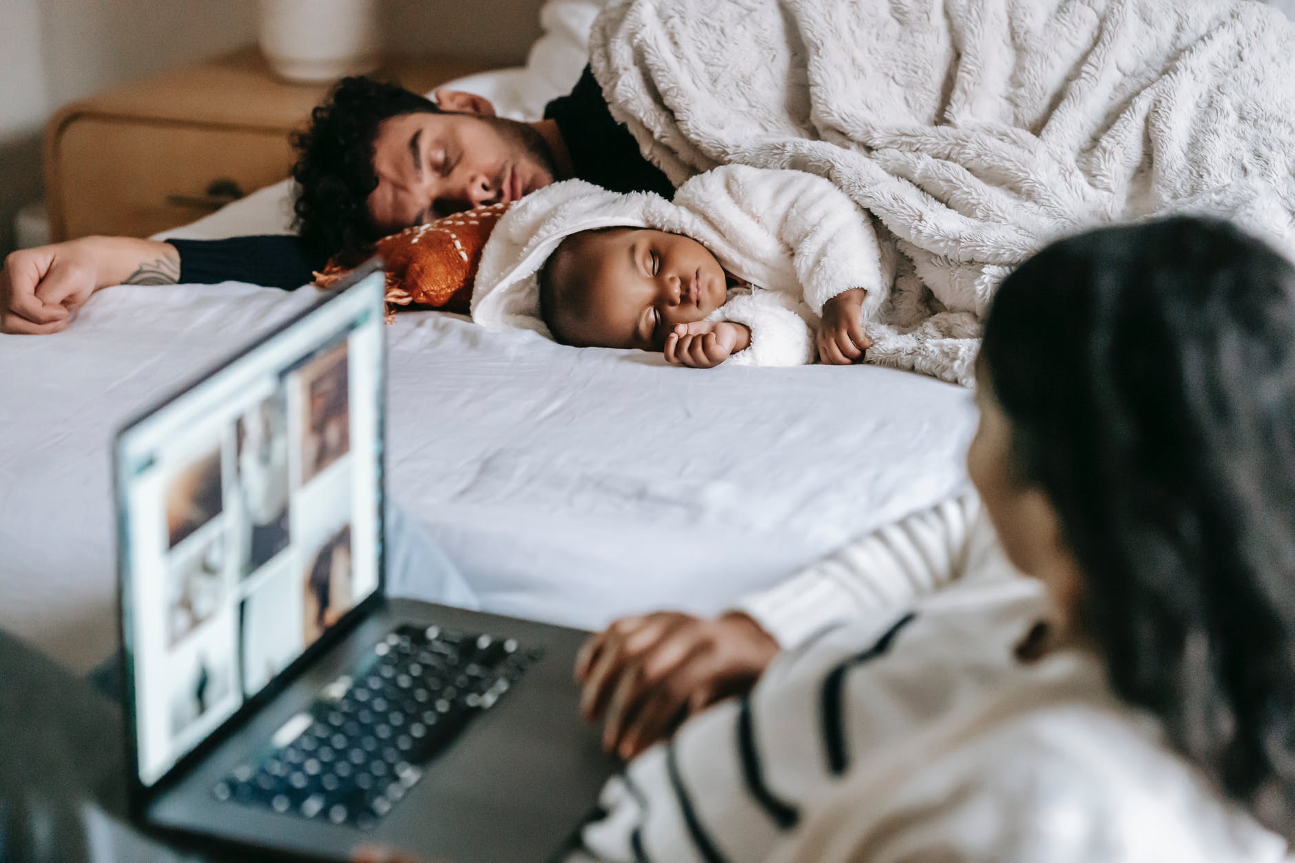 anonymous ethnic mother working on netbook near baby and husband sleeping in bedroom